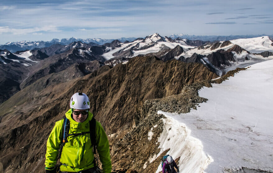 Mann mit grüner Jacke und Helm auf einem Bergrücken. Im Hintergrund mit Schnee bedeckte Gipfelgrate