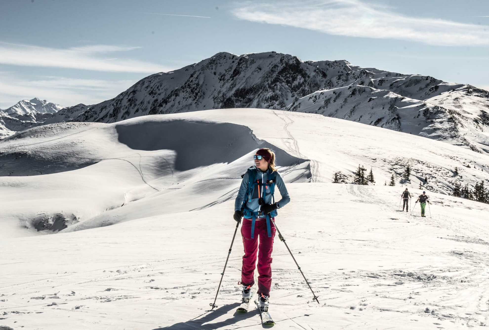 eine hübsche blonde Frau mit Skitourenausrüstung in mitten einer verschneiten Berglandschaft kurz vor dem Gipfel. Im Hintergrund ist eine Wechte zu sehen, die einen Schatten auf die Aufstiegsspur wirft. Es ist ein Sonniger Tag.