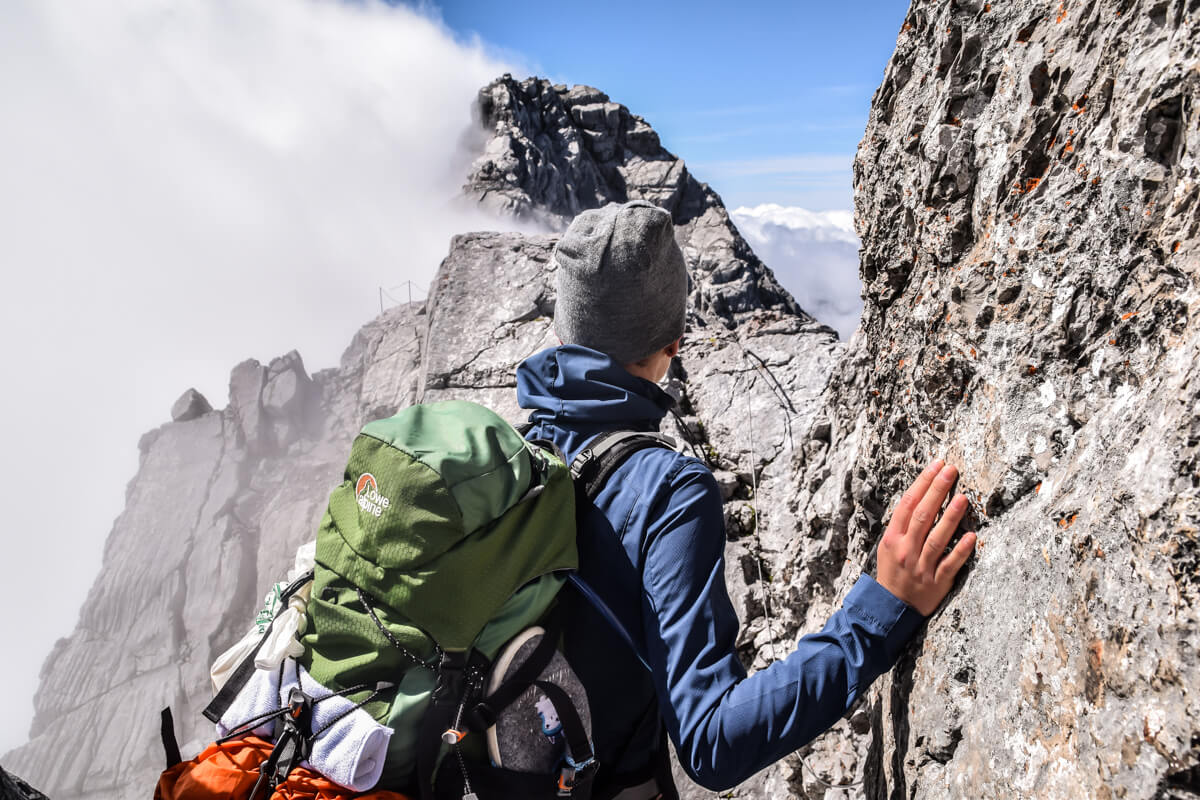 Frau vor einem Zustieg zu einem Alpinen Steig. Im Hintergrund ist ein sehr steiler und ausgesetzter Grad zu sehen