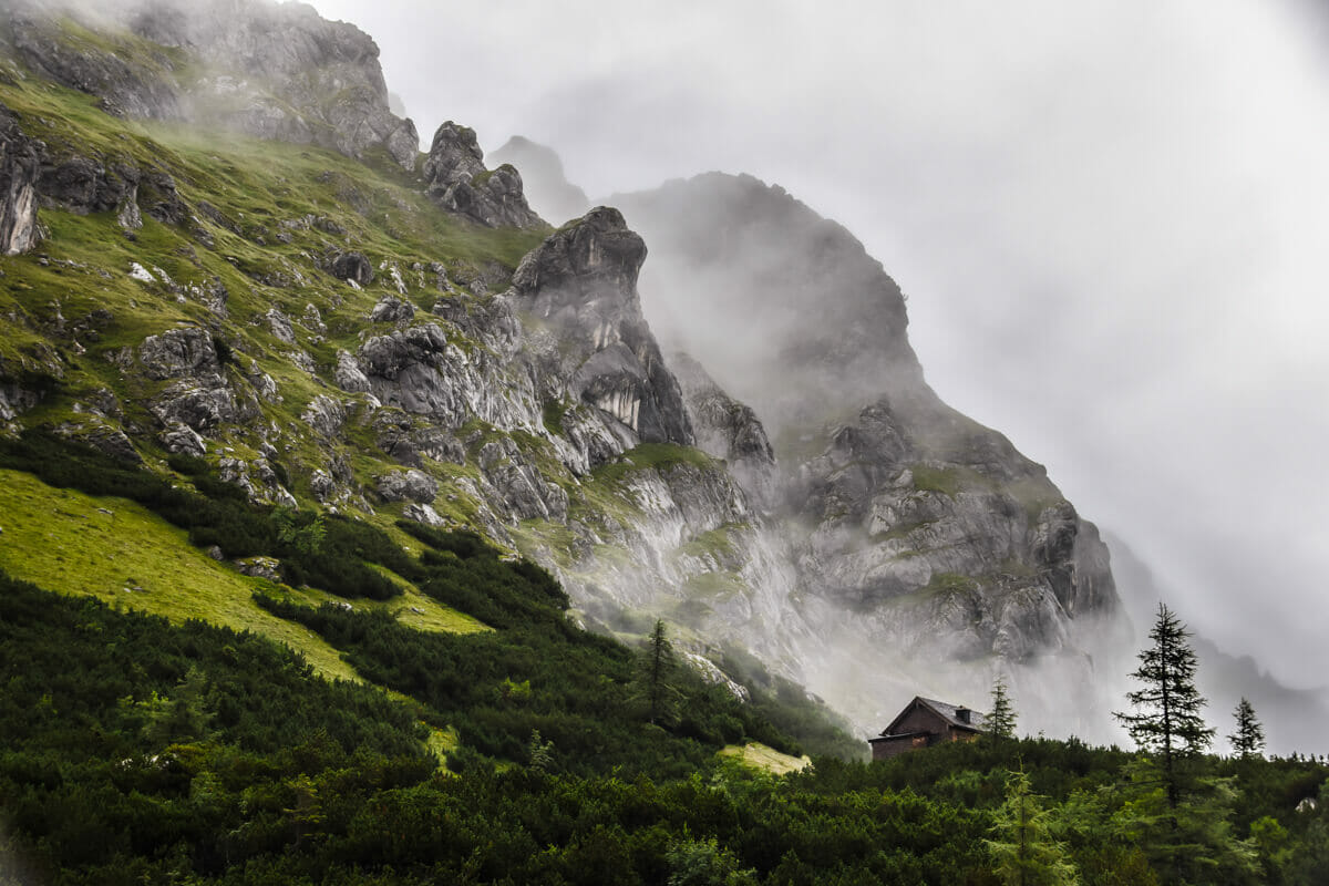 Saftig grüne Berglandschaft im Nebel auf der Watzmanntour. Eine Hütte und Berge sind im Hintergrund zu sehen.