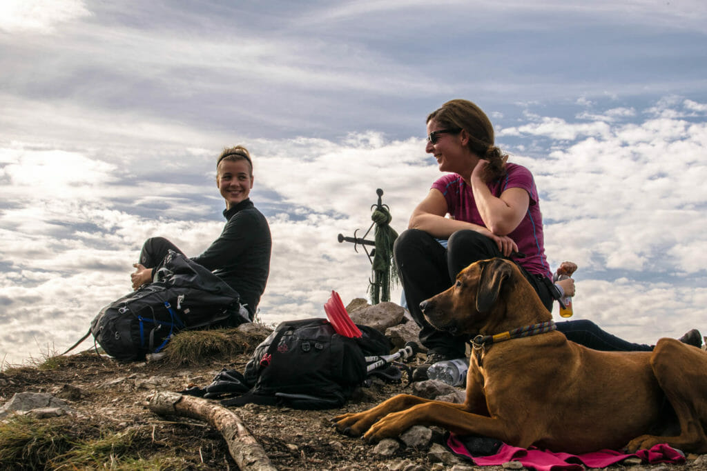 zwei Frauen beim Wandern mit Hund am Gipfel der Sonnenspitze, im Hintergrund Wolken.